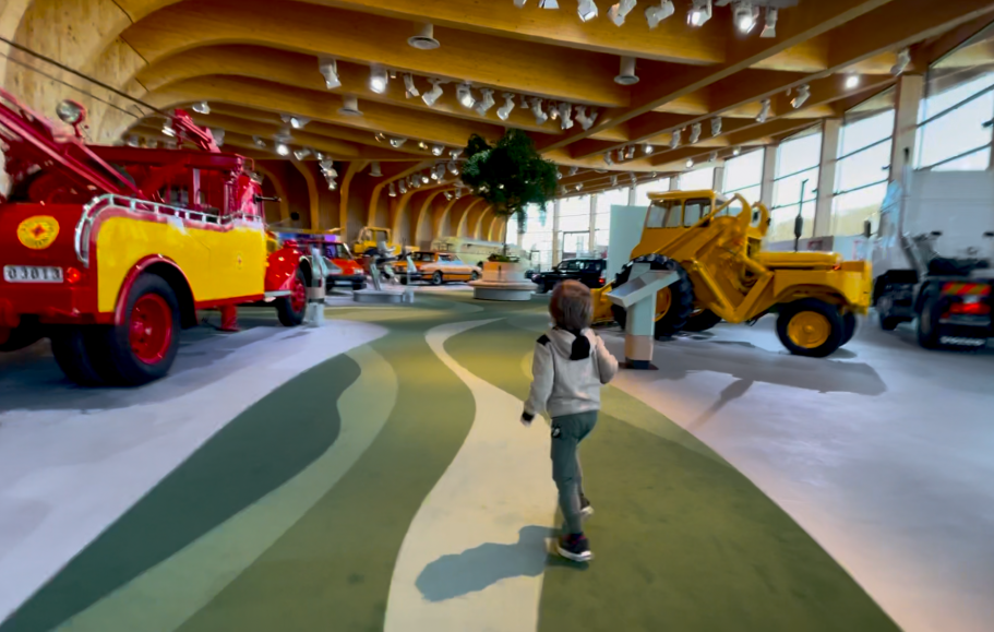 Barn går på en grön matta bland stora maskiner och fordon inuti en modern utställningshall. En gammal röd bärgningsbil, hjullastare, dumper.