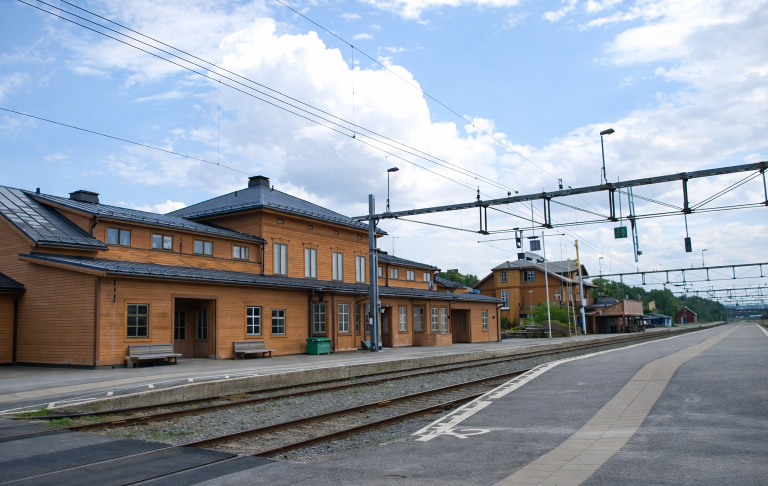 Tågstation med träbyggnader och spår under en blå himmel med moln.