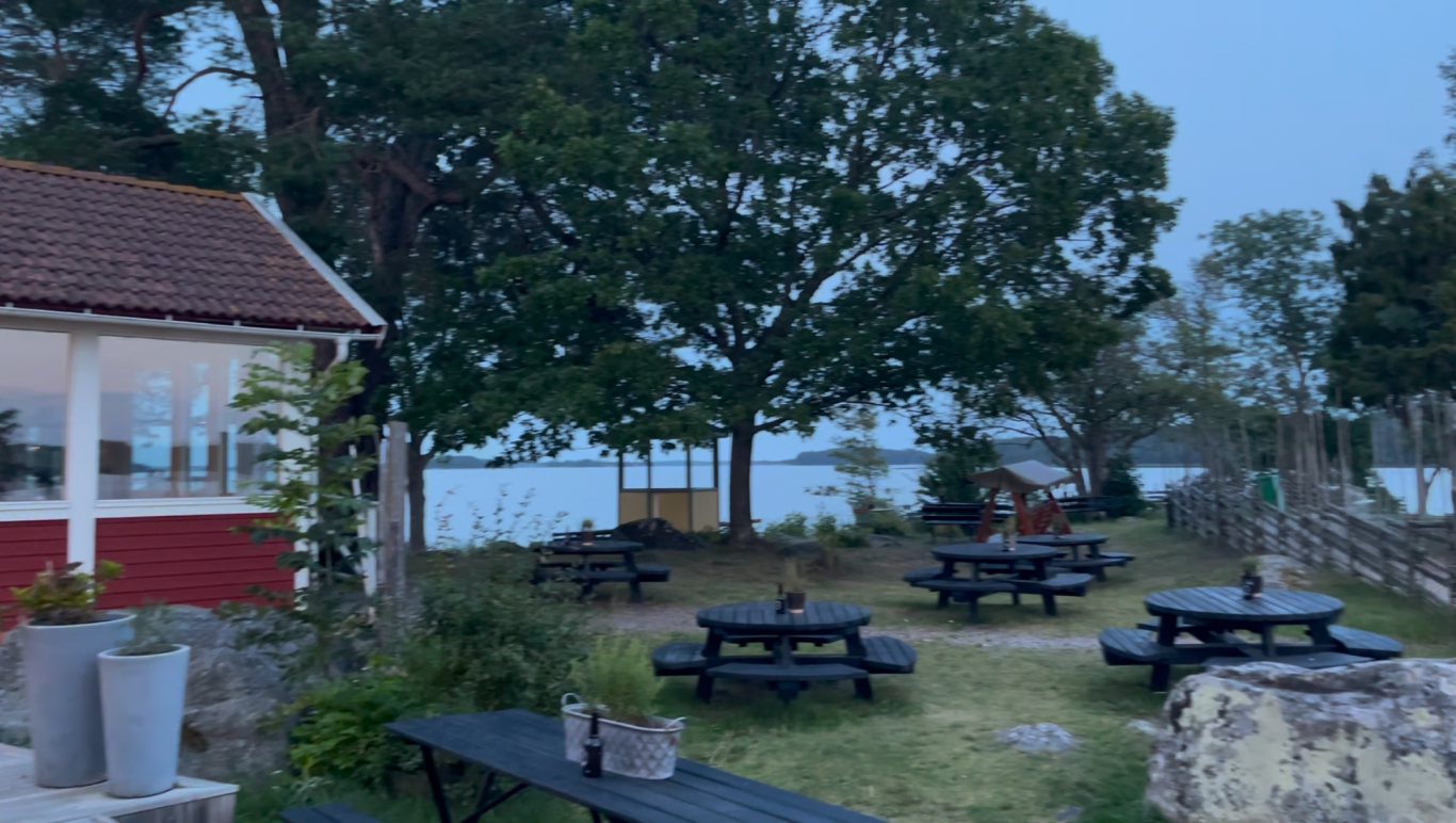 Algots på Udden Uteplats med bänkar och bord vid en sjö, omgiven av träd i skymning.