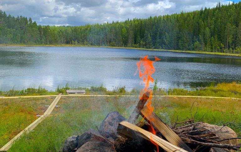 Lugn sjö med omgivande skog och en lägereld i förgrunden.