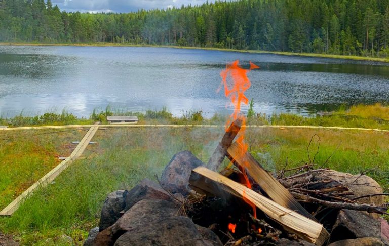 Eldstad vid vindskydd framför sjö Eld i en stenrund bålplats vid en sjö omgiven av grönskande skog.
