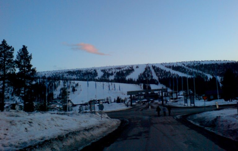 Snötäckt väg med skidbackar i bakgrunden under blå himmel.
