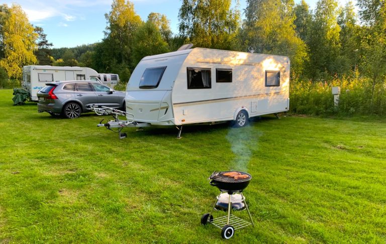 Vit husvagn på camping med en grill som avger rök i grönska. Solnedgången skiner över området.