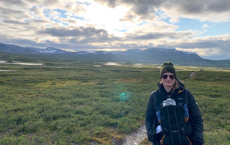 Person i fjällmiljö, bärande mössa och solglasögon, med vidsträckt landskap i bakgrunden.