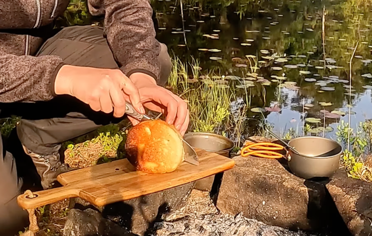 En person skär ett bröd vid en sjö med en gryta bredvid på en träplanka.