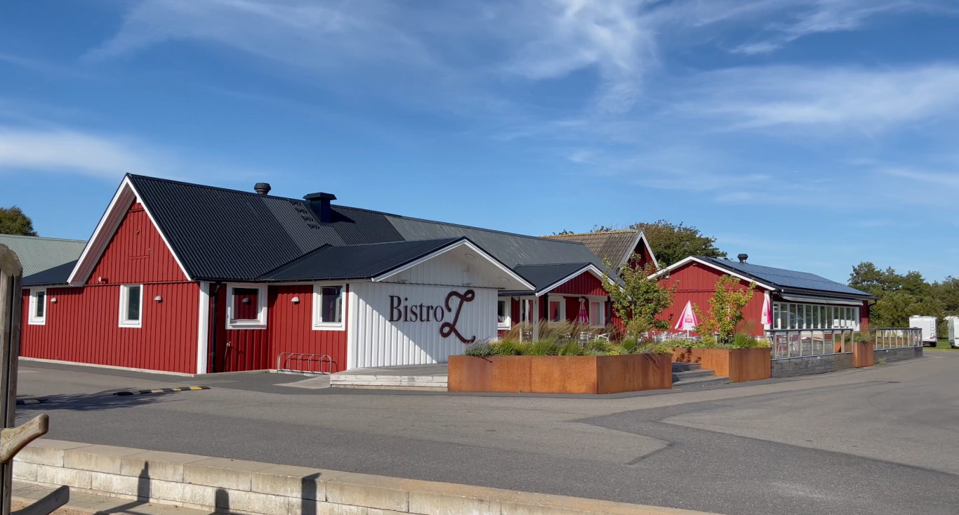 Läjets Camping Äventyrsgolf Röda byggnader med tak i svart, omgivna av grönska och en asfalterad väg.