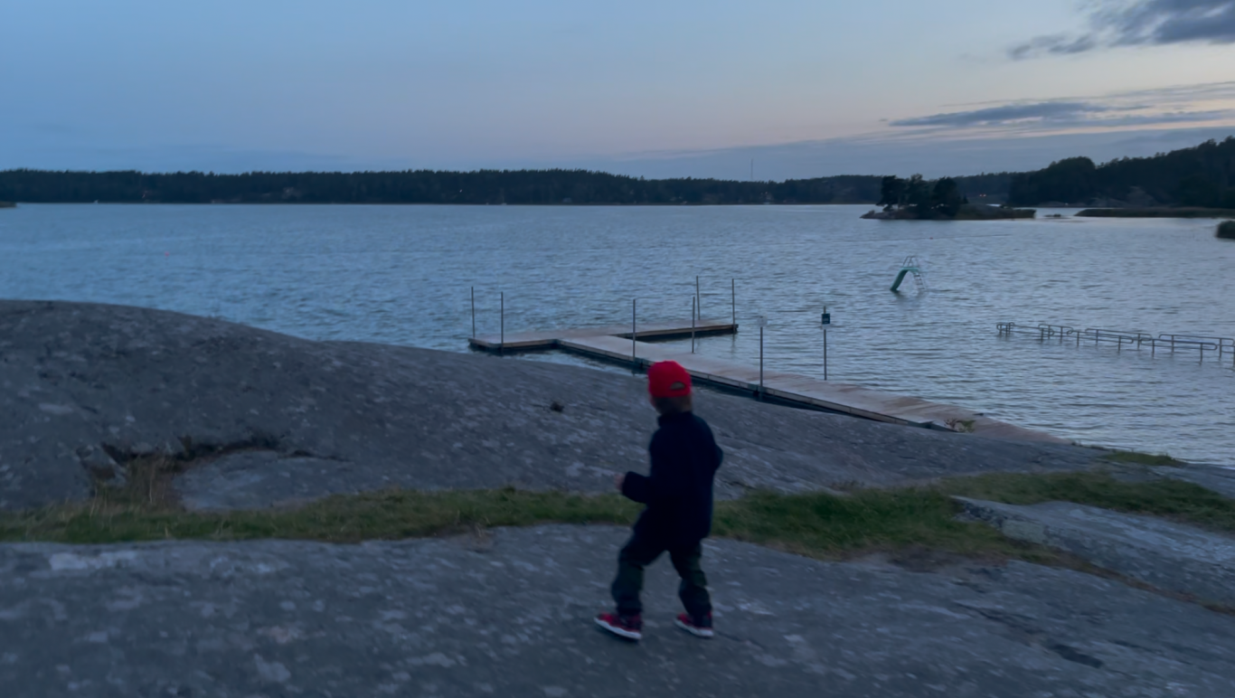 Upptäcker Person i röd mössa går längs en klippa vid vattnet vid skymning.