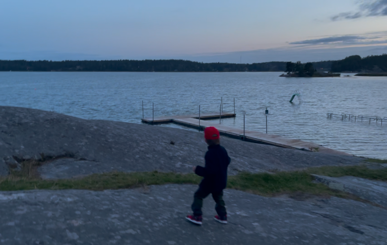 En pojke i röd mössa går på klippor vid en sjö vid skymning.