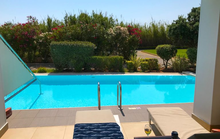 Uteplats med solstolar och en klarblå pool omgiven av grönska.