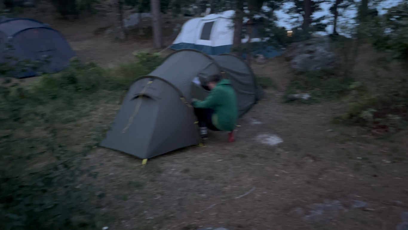 Vår tältplats En person sätter upp ett tält i en skogig campingmiljö.
