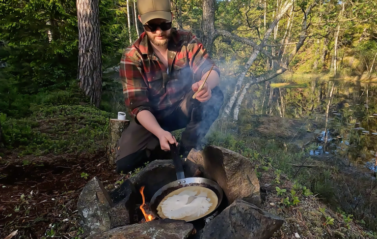 En person lagar mat över öppen eld vid en sjö i skogen. Rök och träd i bakgrunden.
