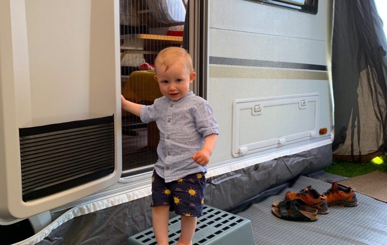 Ett litet barn står vid dörren till en husvagn, iförd blommiga shorts och en t-shirt.