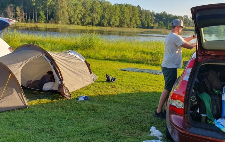 Tält vid sjön med en person som packar bilen och grön gräsmatta runtom.