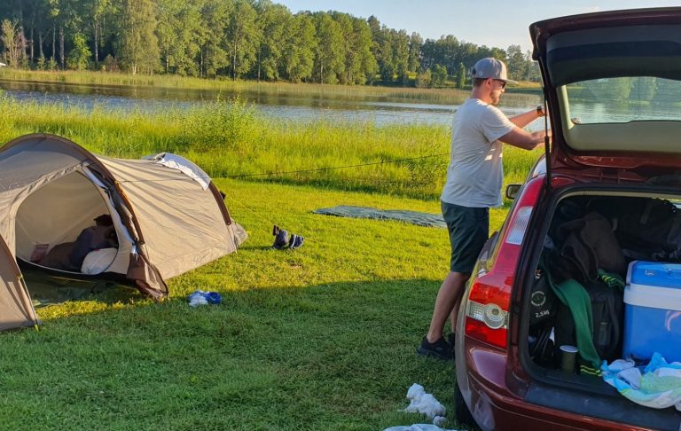 En person packar bilen vid en campingplats vid en sjö, med tält i bakgrunden.