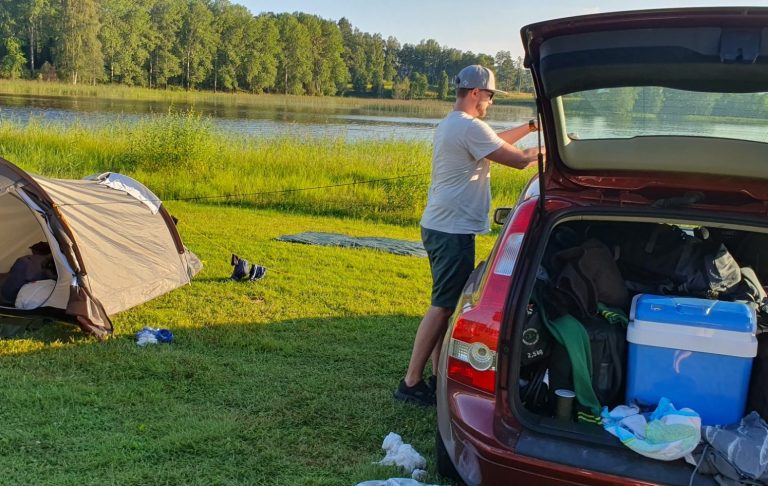 Bilsemester på en Camping En person packar bilen vid en sjö med ett tält och grönska i bakgrunden.