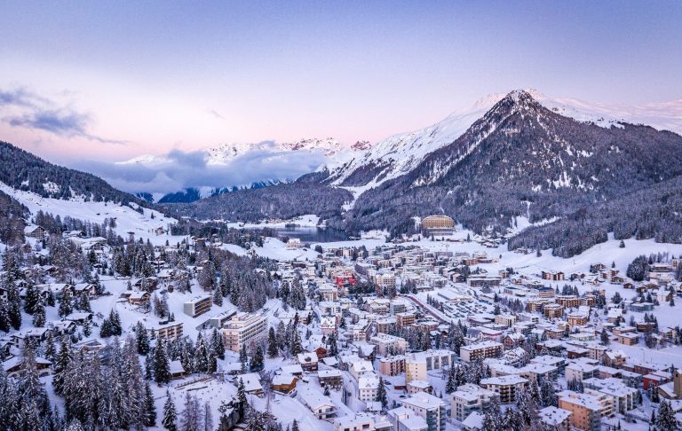 Vinterlandskap med snötäckta berg och en by i dalen under en ljus himmel.