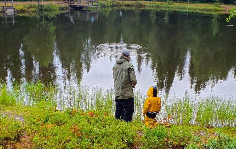 Två personer, en vuxen i grön jacka och ett barn i gul jacka, står vid en sjö.
