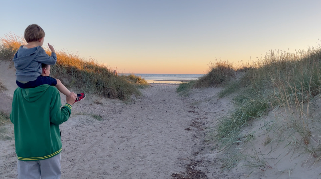 Solnedgång sommarkväll Barn sitter på en vuxens axlar längs en strandstig vid solnedgång.