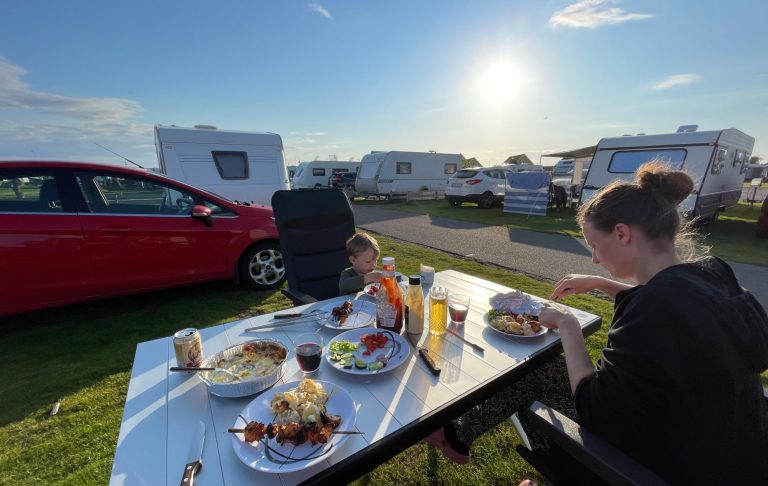 Två personer sitter vid ett bord utomhus med middag, solnedgång och husvagnar i bakgrunden.