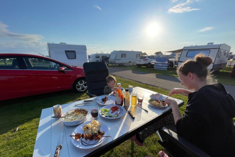Middag på Skrea Camping Två personer sitter vid ett bord med mat, en röd bil och husvagnar i bakgrunden.