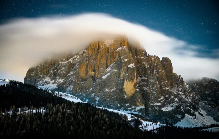 Val Gardena Storslagen bergformation med snö och moln, omgiven av skog och nattlig himmel.