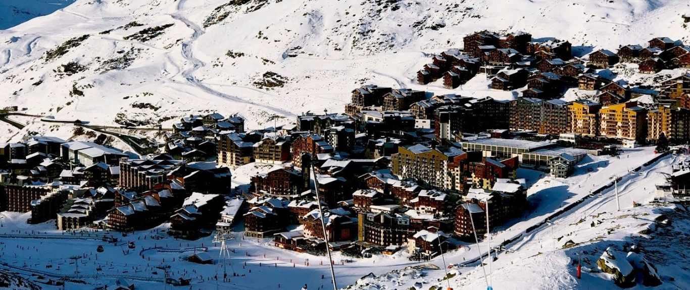 Snötäckt alpby med fjäll i bakgrunden och byggnader spridda över landskapet.