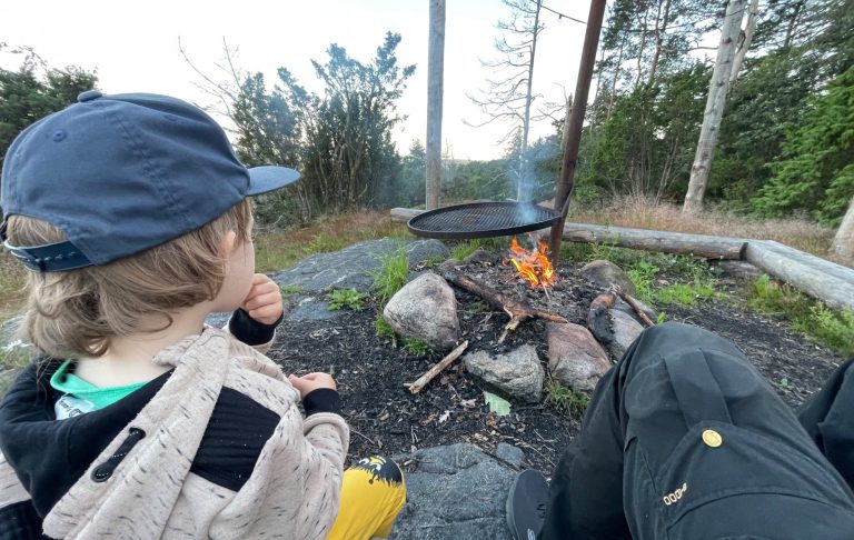 Ett barn sitter vid en eldstad i naturen, tittar på flammorna.