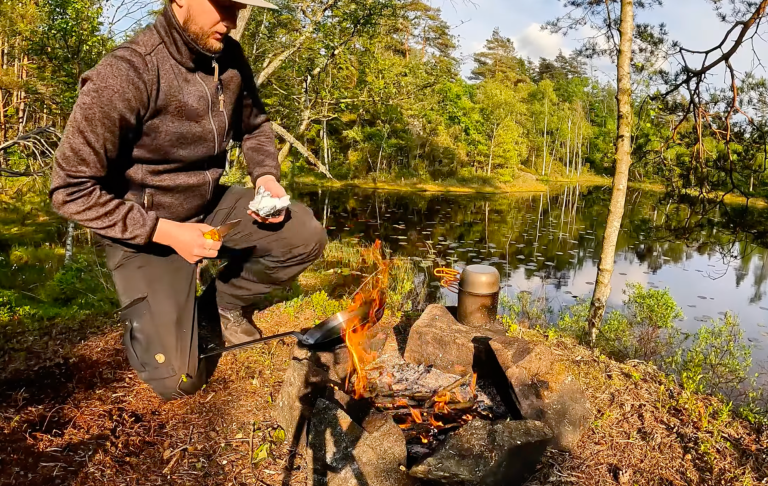 Man lagar mat över en öppen eld vid en sjö, omgiven av grönska.