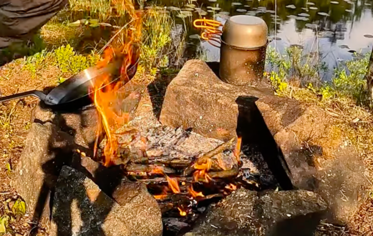 Eld brinner på stenar vid en vattenspegel, med en kastrull över elden.