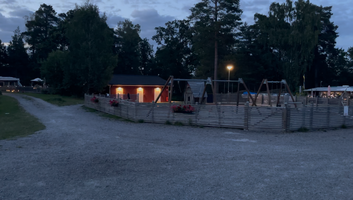 Lekplatsen Ett landskap med en grusväg, stuga och lekplats i skymningens ljus.