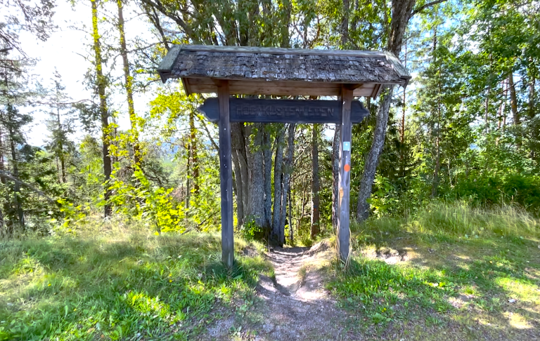 Gammal träportal med tak, omgiven av gröna träd och gräsmatta.