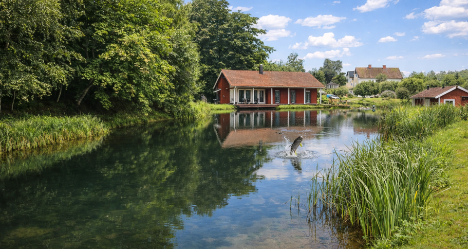 Röd stuga vid en spegelblanka sjö omgiven av grönska och träd.