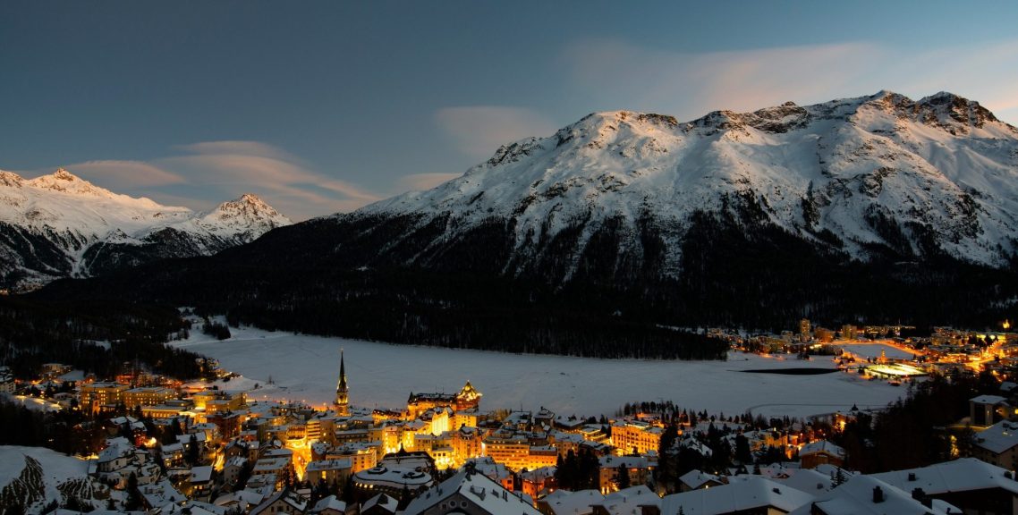 Vy över en snötäckt dal med berg och en belyst alpstad vid en sjö.