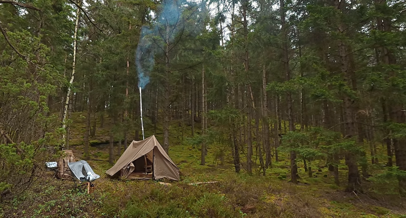 Hot tent Tält i en skog, med rök som stiger från en eldstad. Grönskande omgivning.