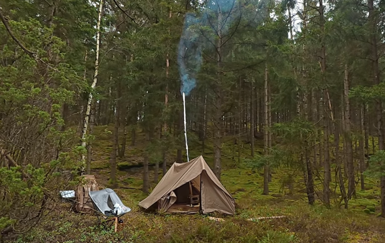Tältkåta i skogen med rök som stiger från skorstenen. Grönska runt omkring.