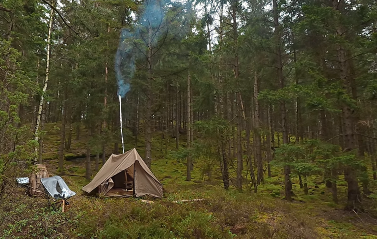 Tält i en skog, med rök som stiger från pipan. Grönskande omgivning.