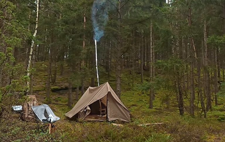 Hot tent med kamin i en skog med rök som stiger från en skorsten, omgiven av träd. Campingstol och ryggsäck står utanför tältet.