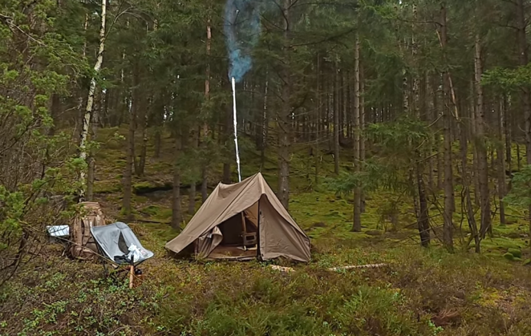 Tält i skog med rök som stiger från en eldstad, omgiven av grön vegetation.