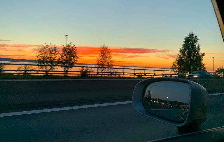 Solnedgång längs vägen Solen går ner över vattnet med träd langs vägen och reflektion i sidomirron.