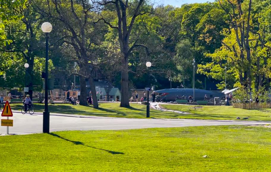 En grön park med träd, belysningspelare och människor som vistas i området. En lekplats syns mitt i bild med barn som leker.