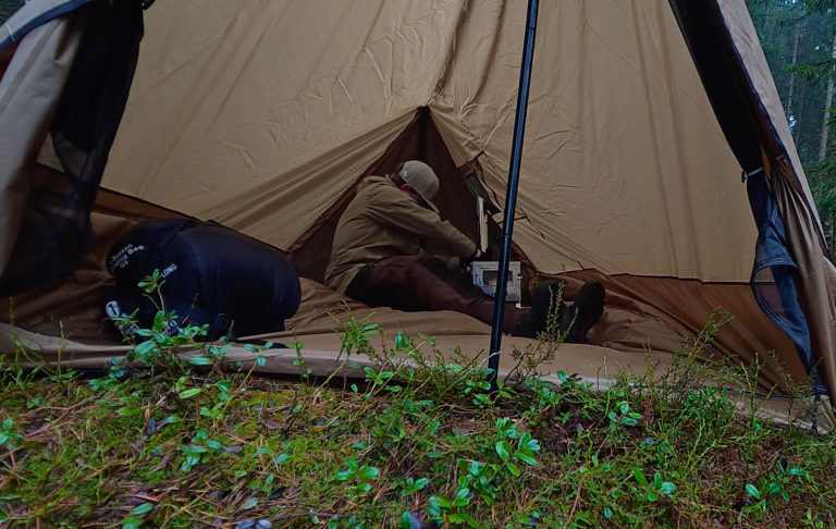 En person sitter inne i ett tält på en campingplats, omgiven av grön vegetation.