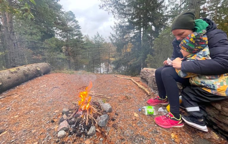 Person sittande vid lägereld i skogen, omgiven av träd och sjö.