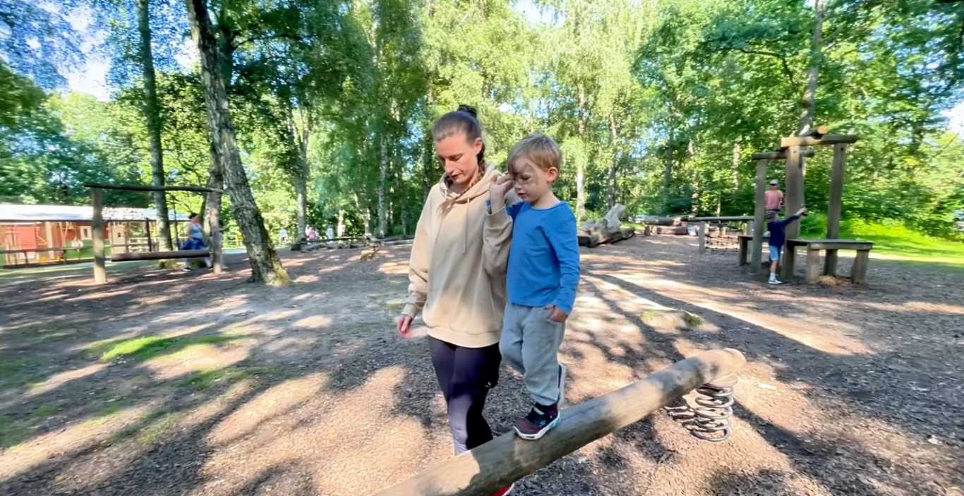 Hinderbanan i Vallarnas Friluftsområde En vuxen leder ett barn som balanserar på en stock i en lekpark omgiven av träd.
