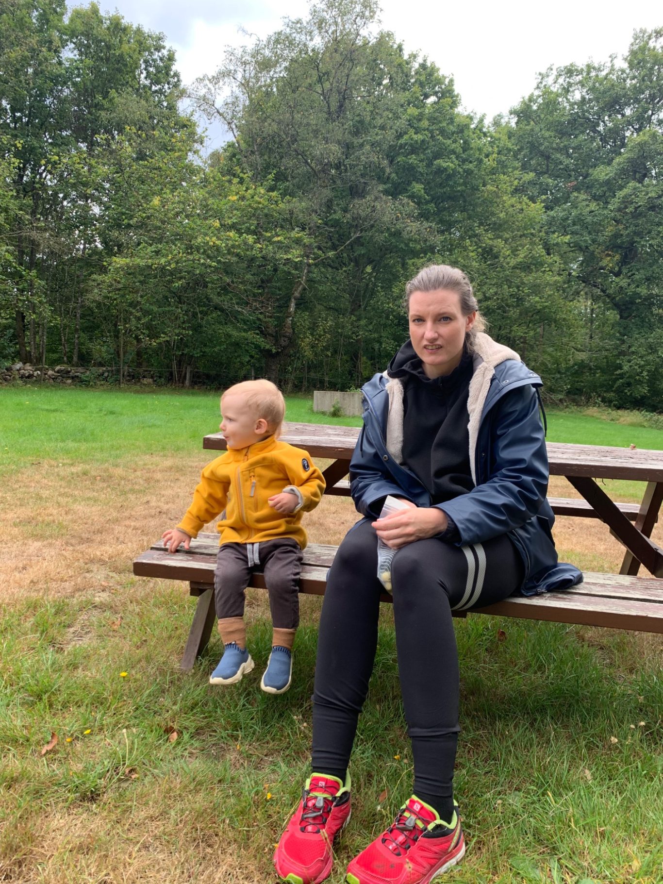 Fikar En vuxen sitter på en bänk med ett litet barn bredvid i en grön park.