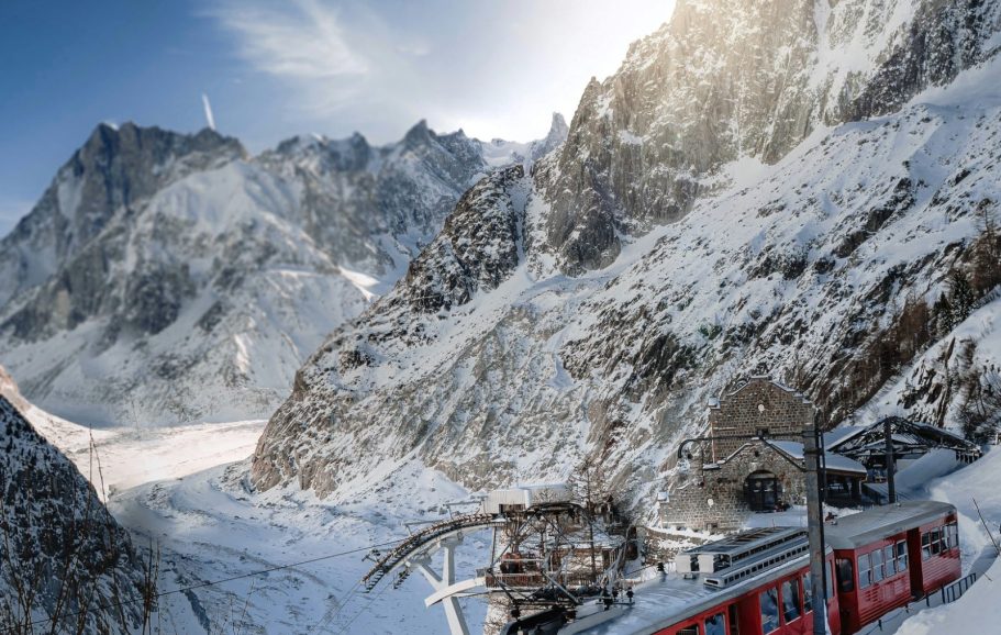 Röd linbana på snötäckta berg med klippiga toppar och klart blå himmel.