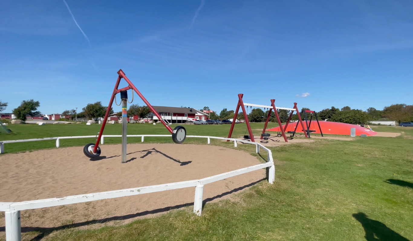 Lekplatsen Björkängs Camping Lekplats med gungor och sandlåda under klart dagsljus. Grön gräsmatta i bakgrunden.