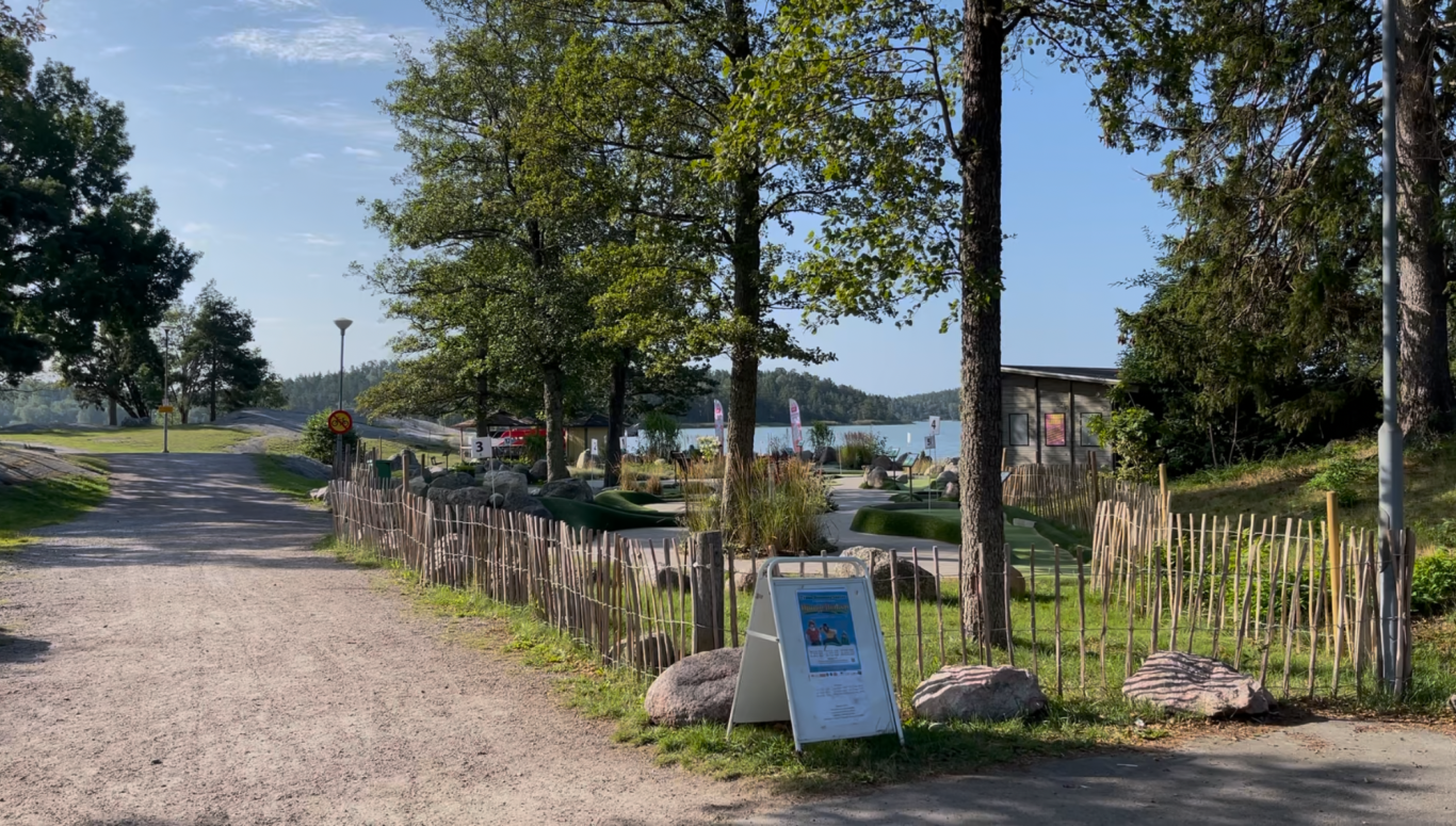 Minigolfbanan vid Stranden Stig vid vattnet med träd och en skylt i förgrunden.