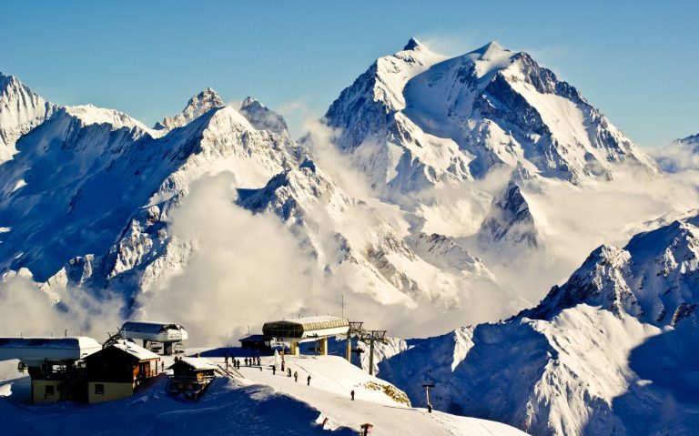 Snöklädda bergtoppar med moln och en fjällstation i förgrunden.