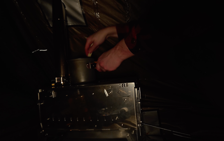 En hand häller vätska från en metallbehållare i en annan behållare. Mörk bakgrund.