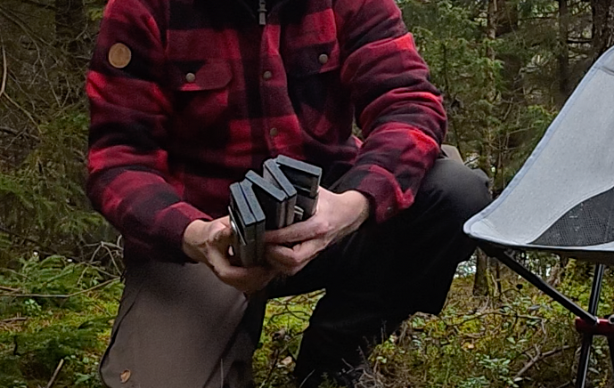 En person i rutig skjorta håller i flera böcker i en skogsmiljö.